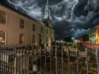 Eerie nighttime view of a historic white church with steeple and adjacent graveyard behind an ornate iron fence, streetlights and light trails under dramatic lightning-filled storm clouds.