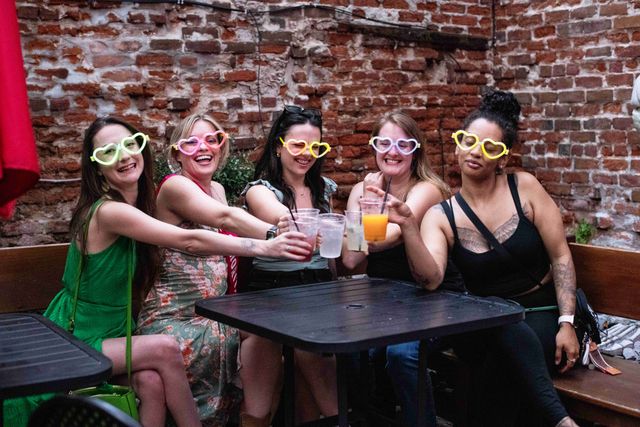 Five friends wearing neon heart-shaped sunglasses toast mixed drinks at a brick-walled outdoor bar patio