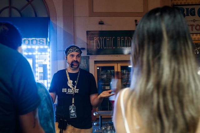 Ghost tour guide wearing a bandana and headset gestures to visitors beneath a glowing “Psychic Reading” sign on a lively downtown night street
