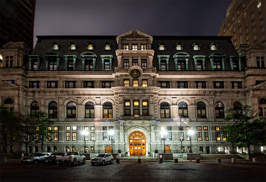 Grand ornate stone government building at night in a downtown plaza — mansard roof with dormers and clock, arched windows, glowing central doorway, streetlamps and parked cars.