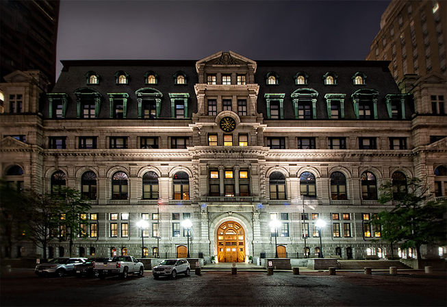 Grand ornate stone government building at night in a downtown plaza — mansard roof with dormers and clock, arched windows, glowing central doorway, streetlamps and parked cars.