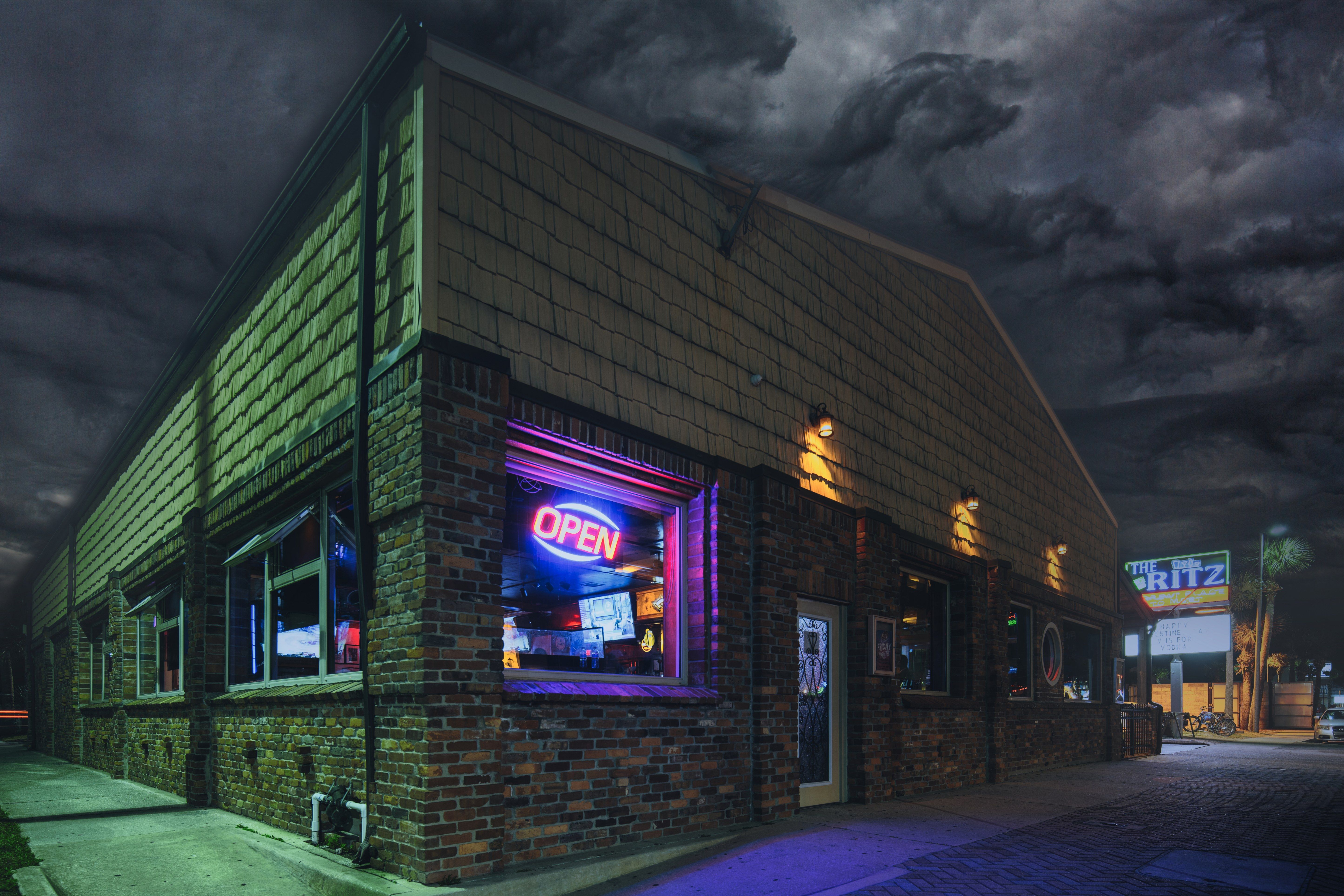 Nighttime urban corner bar in a brick building with a glowing neon OPEN sign in the window, warm exterior lamps, empty sidewalk and moody storm clouds overhead.