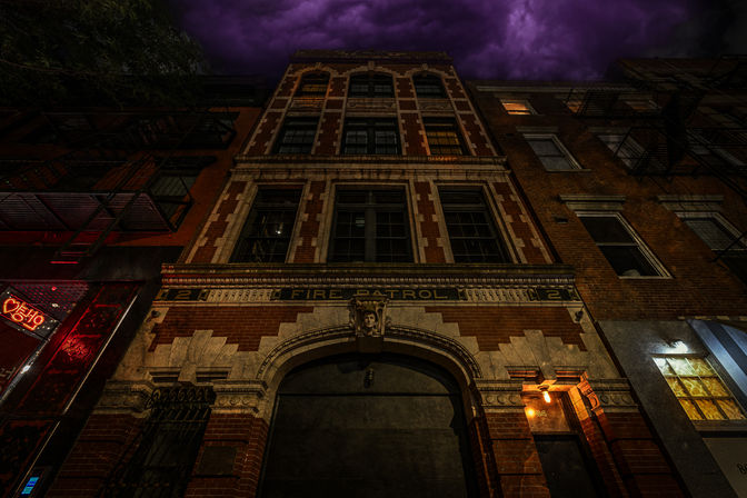 Moody night view looking up at a historic multi-story brick firehouse facade with arched entrance and 'Fire Patrol' lettering, warm amber lighting, adjacent buildings with fire escapes, and a dramatic purple stormy sky overhead.
