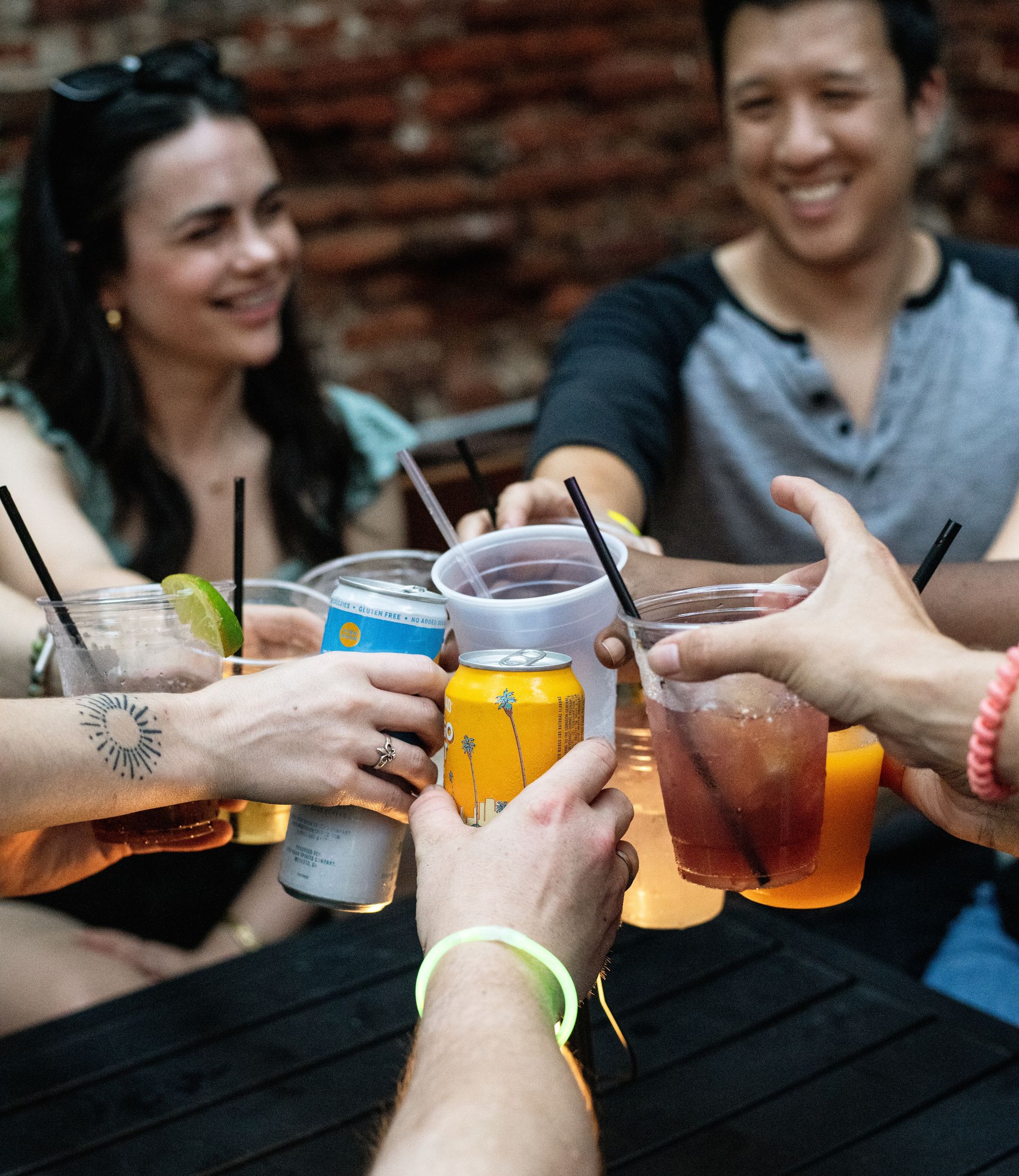 Friends clinking cans and plastic cups of beer and colorful cocktails on an outdoor patio with a brick wall backdrop, casual summer night cheers