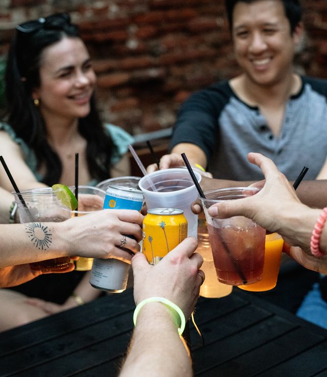 Friends clinking cans and plastic cups of beer and colorful cocktails on an outdoor patio with a brick wall backdrop, casual summer night cheers