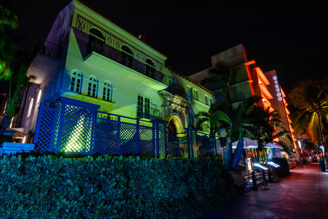 Colorful nighttime scene of an Art Deco–style hotel facade lit in blue, green and red neon, palm trees and a lively tropical sidewalk