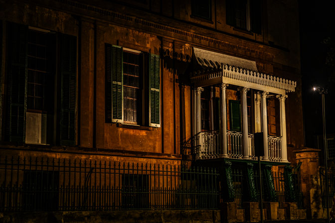 Moody warm-lit historic townhouse facade at night with green shutters, ornate white columned balcony and iron fence.