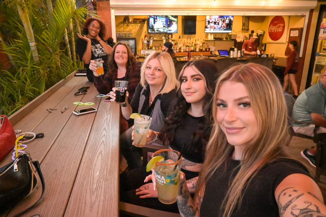 Five friends smiling and holding lime-garnished cocktails at a wooden outdoor bar counter with tropical plants and TV screens in the background.