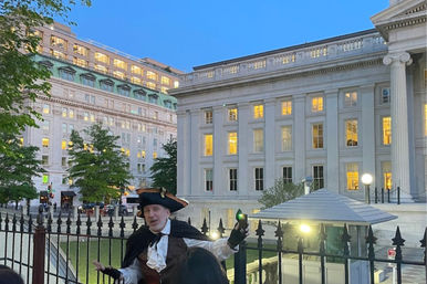 Person in colonial-era costume and tricorn hat holding a lantern by an iron fence in front of a neoclassical government building with glowing windows at dusk in a downtown cityscape.