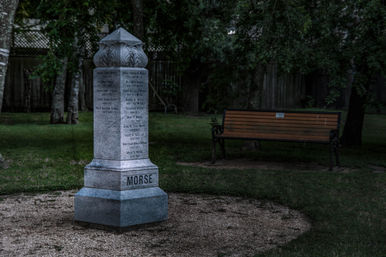 Granite memorial pillar standing in a quiet tree-lined park circle with a wooden bench at dusk.
