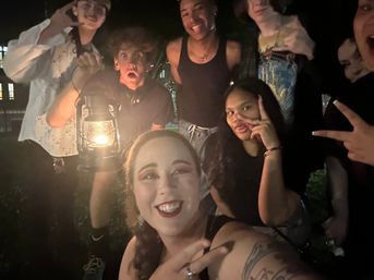 Nighttime outdoor group selfie of friends gathered around a glowing lantern, smiling and striking playful poses and peace signs with warm lantern light highlighting faces and a visible arm tattoo.