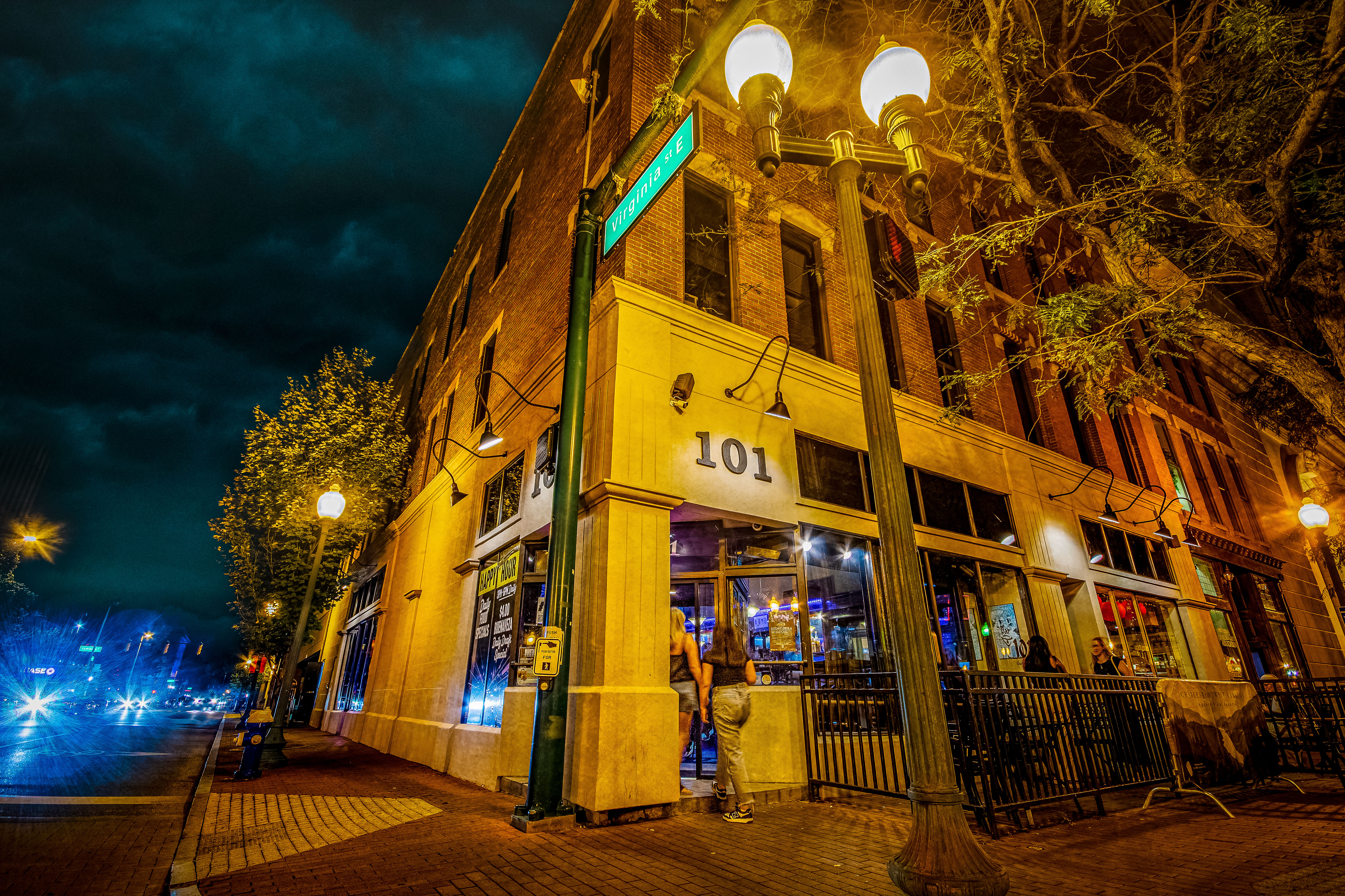 Nighttime urban street corner with a lit brick corner restaurant/bar, glowing street lamps, neon-lit interior and patrons entering under a moody cloudy sky.
