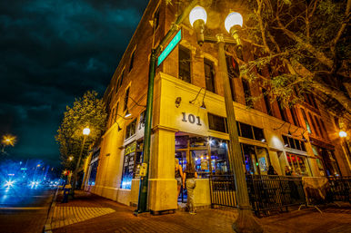 Nighttime urban street corner with a lit brick corner restaurant/bar, glowing street lamps, neon-lit interior and patrons entering under a moody cloudy sky.