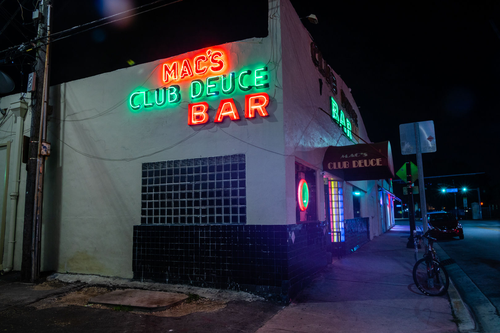 Moody neon-lit corner bar at night with red and green vintage signage, black tile facade and glass-block window, awning, parked bicycle and empty urban sidewalk under streetlights.
