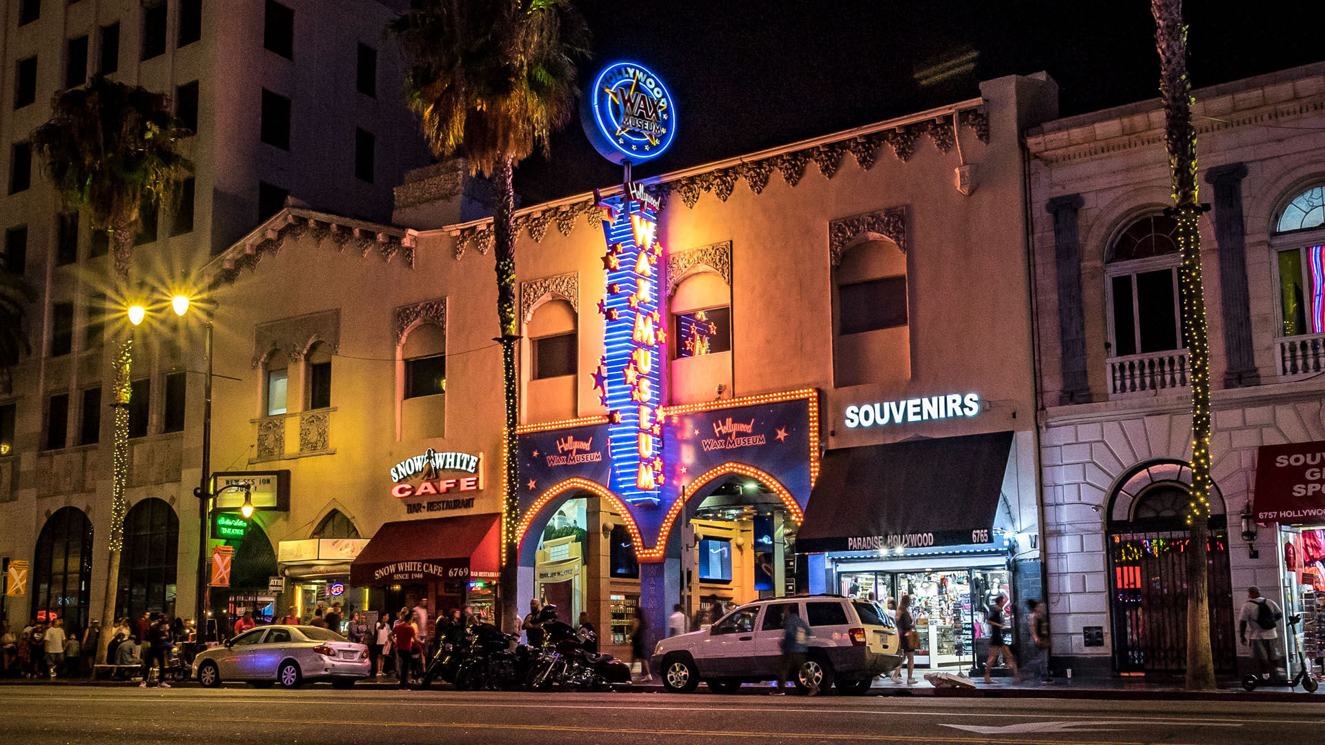 Glowing Hollywood Boulevard at night with a vertical neon wax-museum marquee, souvenir shop awning, palm trees and a bustling sidewalk of pedestrians and parked cars.