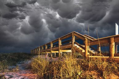 Wooden beach boardwalk over coastal dunes and tall grasses, warmly lit against looming dark mammatus storm clouds for a dramatic, moody coastal scene.