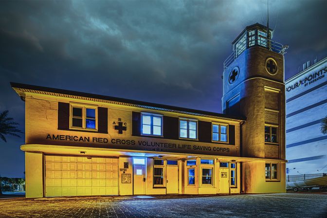 Historic beachfront volunteer life-saving station with tower and visible cross emblems, glowing golden and blue under a dramatic stormy night sky and empty paved forecourt.