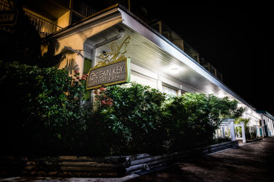 Nighttime view of a lit coastal resort entrance with a wooden hanging sign, lush shrubs, and a white covered porch walkway.