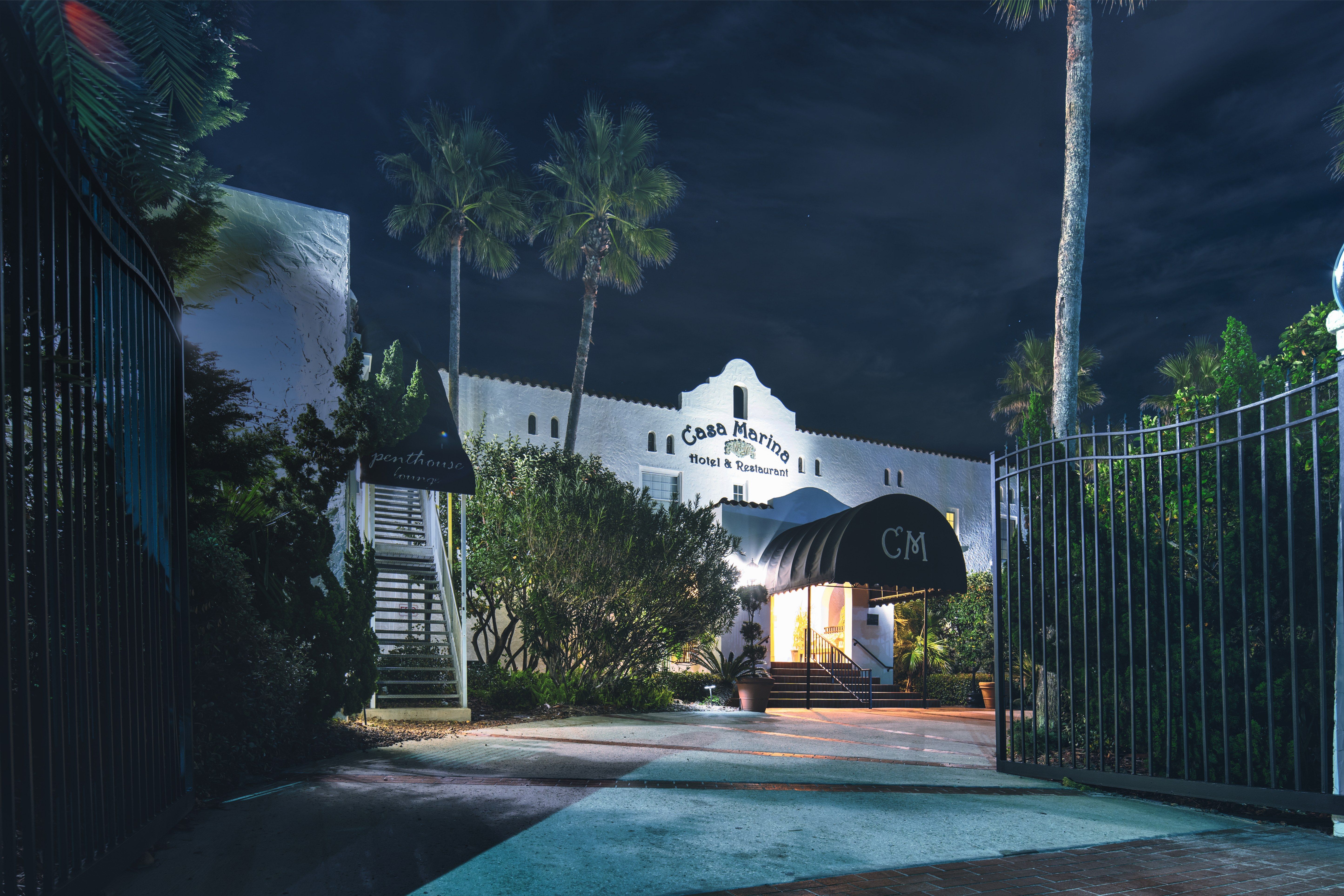 Nighttime view of a white Mediterranean-style coastal hotel entrance with an illuminated arched canopy, palm trees, and an open wrought-iron gate.