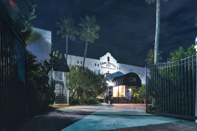 Nighttime view of a white Mediterranean-style coastal hotel entrance with an illuminated arched canopy, palm trees, and an open wrought-iron gate.