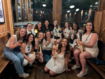 Group of a dozen women smiling and posing inside a rustic wooden bar, many dressed in white, holding cocktails and novelty wooden paddle cutouts with printed photos while sitting on benches and enjoying a lively night out.