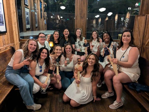 Group of a dozen women smiling and posing inside a rustic wooden bar, many dressed in white, holding cocktails and novelty wooden paddle cutouts with printed photos while sitting on benches and enjoying a lively night out.