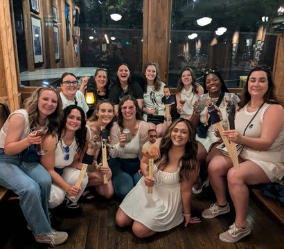 Smiling group of women at a rustic wood-paneled bar, holding cocktails and novelty paddle cutouts for a lively girls' night out celebration
