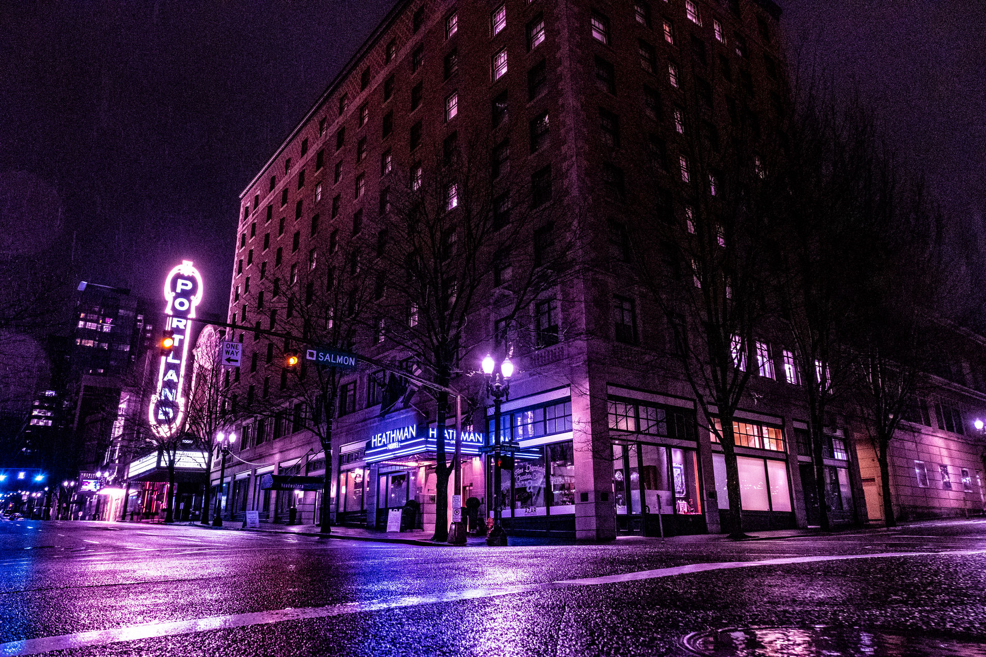 Neon-lit night in downtown Portland: a tall brick corner building and glowing vertical sign cast purple-pink reflections on a rain-slick street.