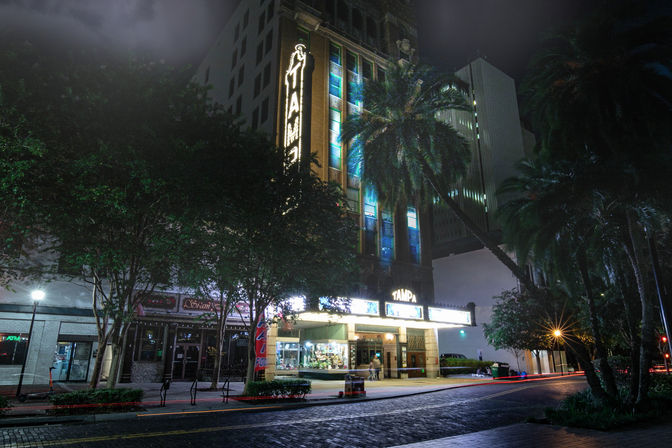 Downtown Tampa nighttime scene with a historic theater marquee glowing 'Tampa', tall palm trees, wet brick street and illuminated storefronts.
