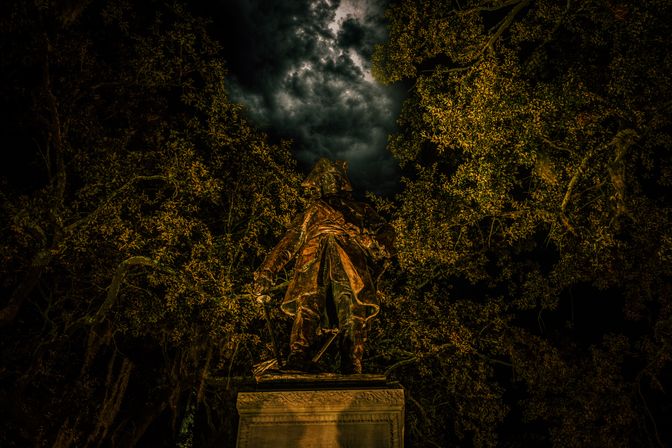 Nighttime bronze statue of a cloaked historical figure on a pedestal, framed by oak trees and dramatic moonlit clouds