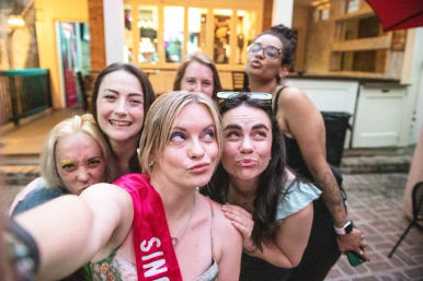 Playful group selfie of six friends on a brick restaurant patio at dusk, a celebrant wearing a pink sash and the group making silly faces and poses.