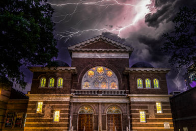 Historic downtown brick church facade at night with glowing stained-glass windows and ornate arched doors, dramatic lightning streaking across a purple storm sky, framed by trees and nearby urban buildings.