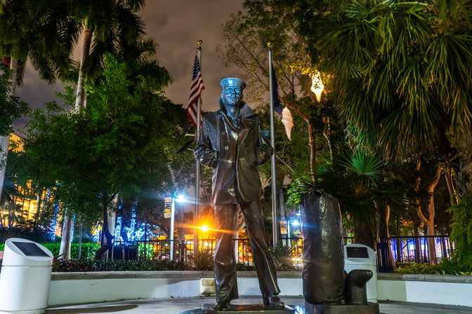 Bronze statue of a sailor in a pea coat and cap standing next to a duffel bag, lit at night in a palm‑lined plaza with an American flag and colorful city lights in the background.