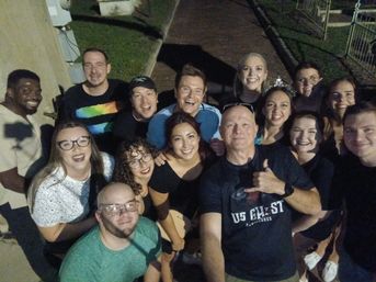 Large outdoor nighttime group selfie of smiling friends huddled on a brick walkway, casual clothes and playful expressions