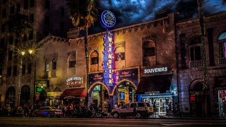 Vibrant Hollywood nighttime street scene with a neon museum marquee, souvenir shop and café, palm trees and crowds under dramatic clouds.