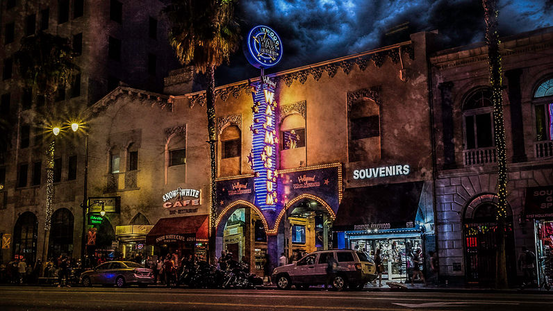 Vibrant Hollywood nighttime street scene with a neon museum marquee, souvenir shop and café, palm trees and crowds under dramatic clouds.