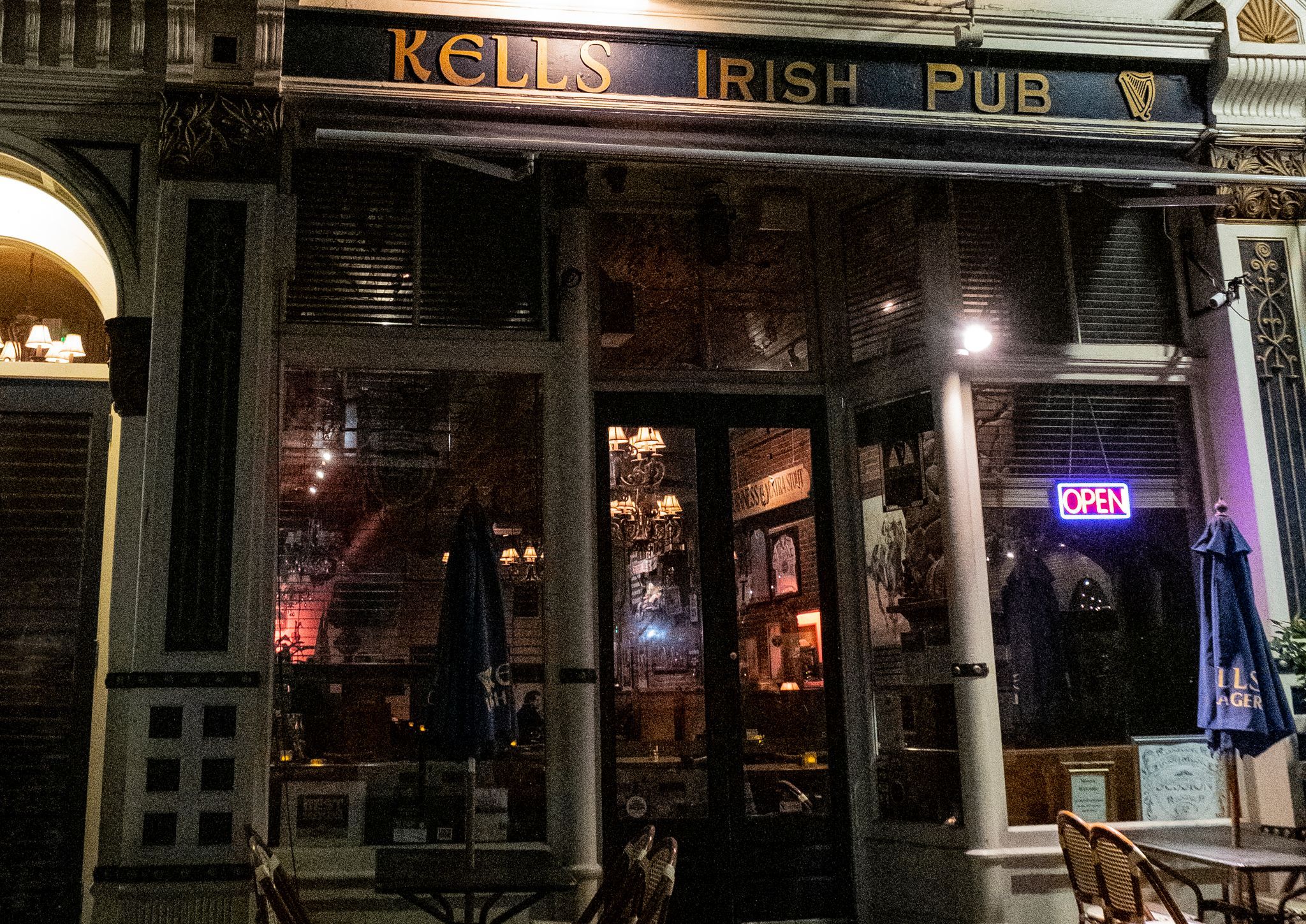 Nighttime Irish pub storefront on a city street, classic dark facade with illuminated "OPEN" neon sign, outdoor bistro tables and umbrellas and warm chandelier glow through glass doors.
