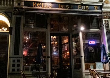 Nighttime Irish pub storefront on a city street, classic dark facade with illuminated "OPEN" neon sign, outdoor bistro tables and umbrellas and warm chandelier glow through glass doors.