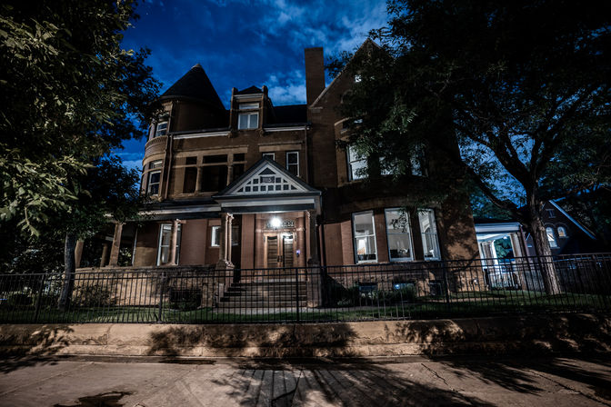 Historic brick Victorian mansion at night with a turret, illuminated front porch and bay windows, set behind a wrought-iron fence and framed by trees under a deep blue sky.