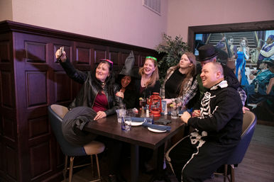 Six friends in costume gathered at an indoor restaurant booth — witch hat, skeleton onesie and top hat visible — laughing and taking a selfie over a table with drinks and a red lantern.