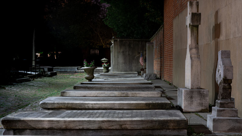 Moody night view of a historic cemetery with stone tomb slabs lined along a brick wall, upright headstones, planters, and an American flag