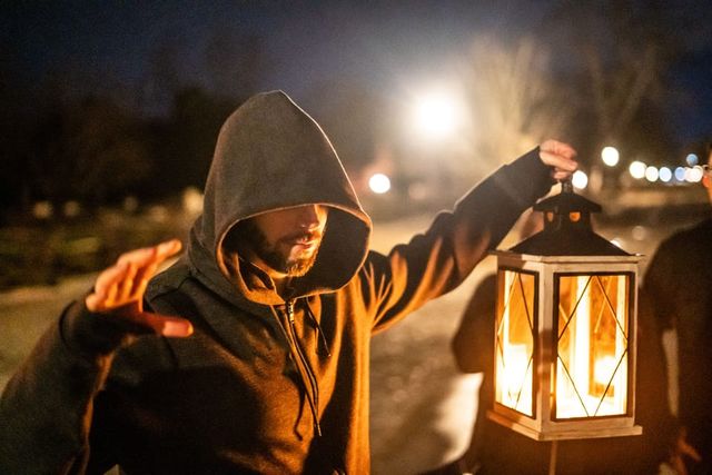 Hooded person holding a glowing lantern on a dim nighttime path, warm light illuminating their face with bokeh streetlights in the background.
