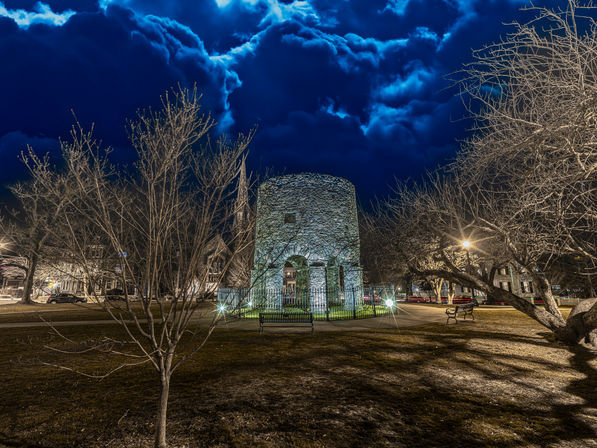 Historic round stone tower in a small town park at night, illuminated by lamp posts and spotlights, surrounded by leafless trees and benches beneath dramatic deep-blue storm clouds.