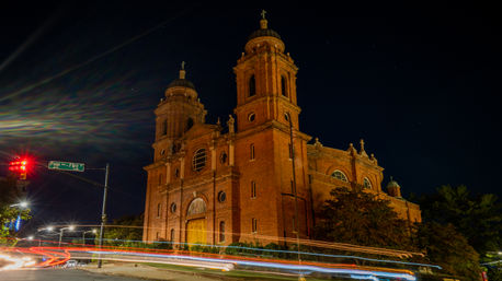 Illuminated red-brick church with twin towers at night, dynamic long-exposure car light trails streaking across the intersection and a glowing traffic light under a deep navy sky.