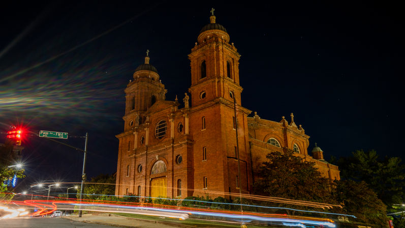 Illuminated red-brick church with twin towers at night, dynamic long-exposure car light trails streaking across the intersection and a glowing traffic light under a deep navy sky.