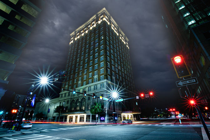 Downtown night scene: illuminated brick high-rise at a busy intersection under stormy clouds, starburst streetlights and red signals with car light trails.
