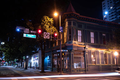 Nighttime Pine St corner with an ornate Victorian-style building bathed in warm streetlight, red traffic signals and parking sign, tree-lined sidewalk and streaking car light trails.