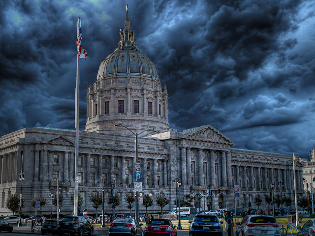 Dramatic storm clouds over a Beaux-Arts domed city hall with an American flag and parked cars in downtown San Francisco