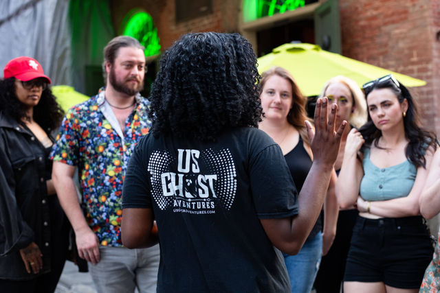 Person in a black t-shirt leading a ghost walking tour, gesturing to a small group of adults on a brick-lined city street with neon green arched windows and a bright yellow umbrella
