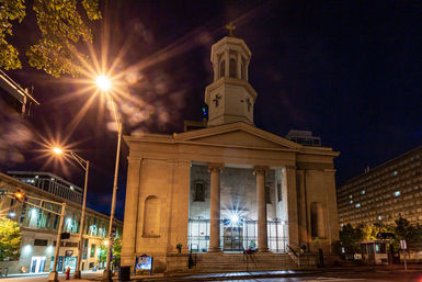 Neoclassical church with tall columns and an illuminated steeple topped by a cross on a downtown city street at night, glowing streetlights creating starburst light effects.